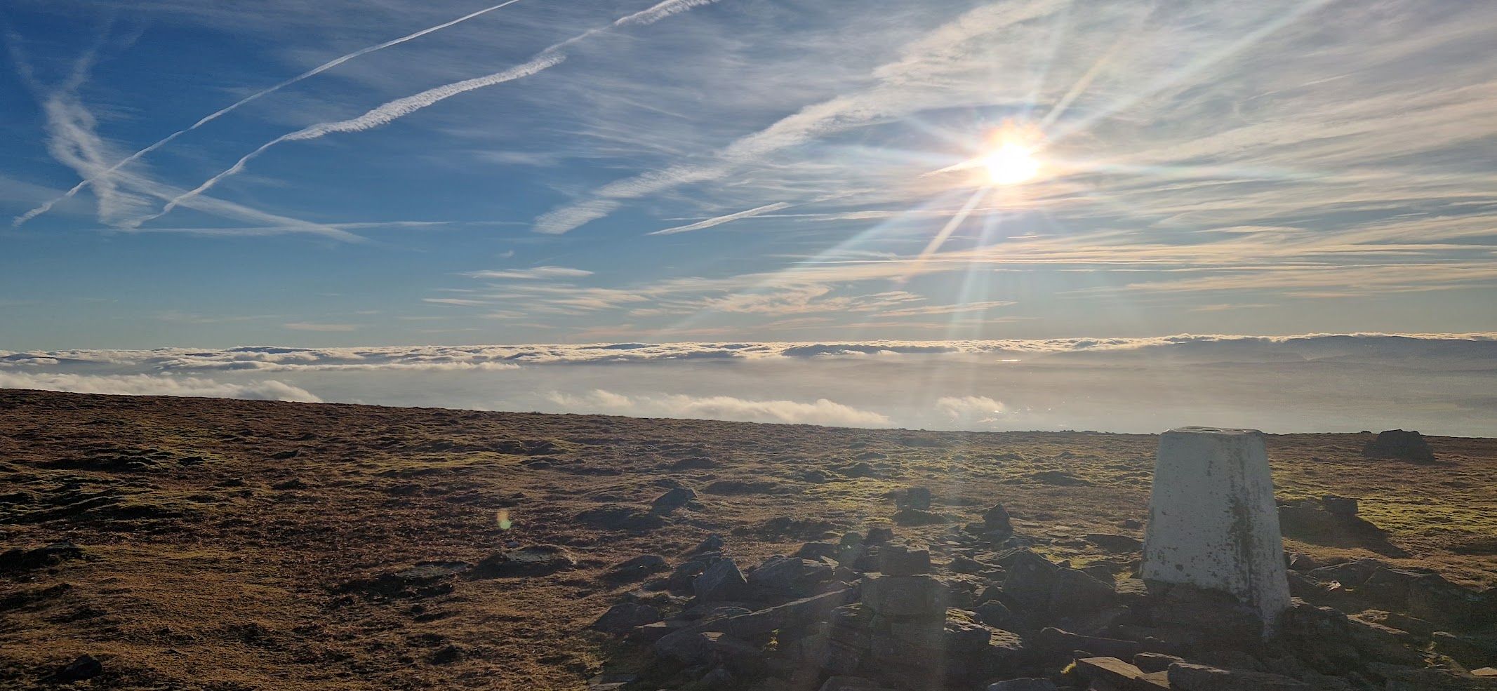 A summit trig point on open moorland, above a sea of cloud